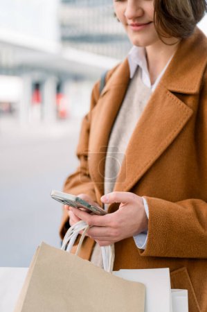 Photo for Crop of Young woman with paper bag and smartphone smiling and looking at screen while standing on blurred background of city after shopping day - Royalty Free Image