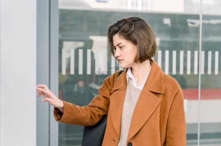 Photo for Side view of young woman with brown short hair in coat checking timetable at bus stop and waiting for transportation after work - Royalty Free Image