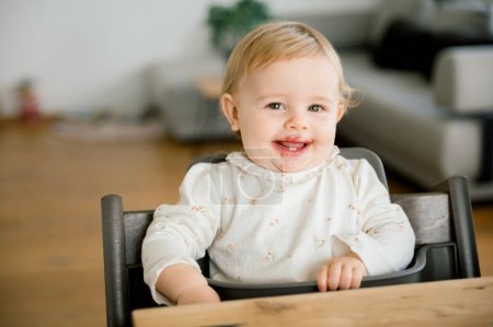 Photo for Baby girl wearing knitted sweater sitting in high chair and feels hungry, waiting fresh fruit puree or porridge, looking at camera and smiling, background living room. family concept - Royalty Free Image