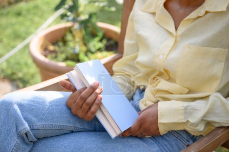 Téléchargez les photos : Récolte de mains féminines tenant un livre pendant qu'elle se relaxe sur une chaise par une journée ensoleillée dans le jardin, les plantes et les fleurs en arrière-plan - en image libre de droit