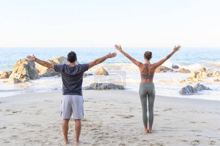 Foto de Vista trasera de un hombre irreconocible en ropa deportiva parado en la playa de arena y estirando los brazos mientras practica yoga en un día soleado - Imagen libre de derechos