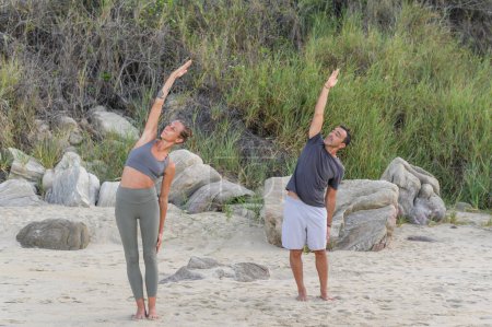 Téléchargez les photos : Corps complet de jeune homme en vêtements de sport debout avec les bras levés et les yeux fermés tout en pratiquant le yoga sur une plage de sable fin - en image libre de droit