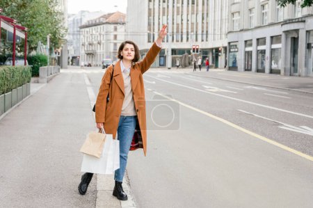 Photo for Full body of young female in casual clothes, coat, jeans, after work standing on asphalt road, holding shopping bags and waving taxi, in background urban scene - Royalty Free Image