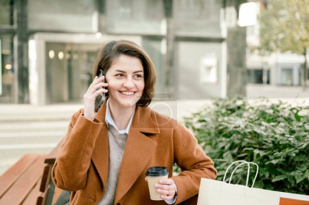 Photo for Positive young female in casual clothes smiling, sitting on bench while taking a break, while having conversation on mobile phone and drinking coffee - Royalty Free Image