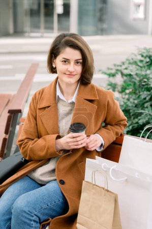 Foto de Joven hembra positiva en traje de moda sonriendo y mirando a la cámara mientras está sentado en el banco con una bolsa de papel y sosteniendo una taza de café - Imagen libre de derechos