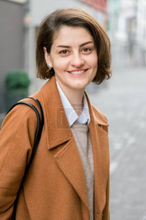 Photo for Positive young female with brown short hair, wearing earrings in elegant coat smiling and looking at camera while standing on city street - Royalty Free Image