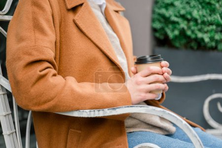 Foto de Vista lateral de la cosecha irreconocible hembra en ropa casual, abrigo, jeans sentados en el banco y sosteniendo la taza de bebida caliente mientras toma un descanso de la oficina - Imagen libre de derechos