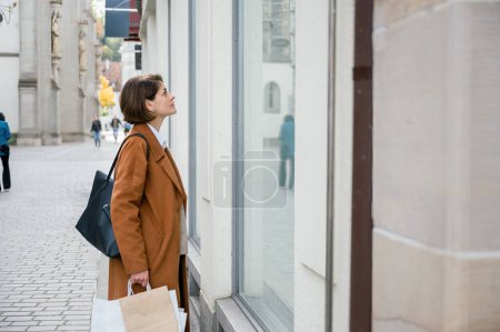 Photo for Side view of young female in smart casual clothes, coat, jeans, handbag standing on city street and looking shop window while shopping in town. - Royalty Free Image
