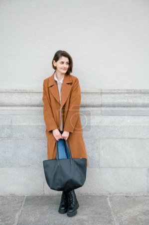 Téléchargez les photos : Corps complet de jeune femme confiante dans un manteau élégant avec sac à main debout contre un mur gris et regardant la caméra - en image libre de droit