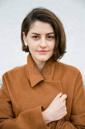 Photo for Attractive young female with short hair, wearing earrings, in brown coat looking at camera on white background - Royalty Free Image