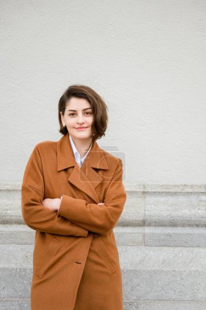 Photo for Confident young female entrepreneur in trendy coat and with arms crossed looking at camera while standing near gray wall on city street, copy space, fashion concept - Royalty Free Image