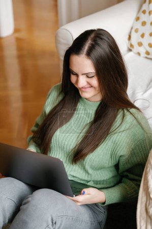 Téléchargez les photos : Jeune femme concentrée aux longs cheveux bruns, vêtue de vêtements décontractés, travaillant sur un ordinateur portable, assise confortablement sur le sol dans un environnement confortable à la maison - en image libre de droit