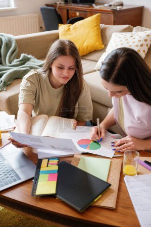 Photo for Two young women, aged 20-22, engaged in focused studying together in a comfortable apartment setting, epitomizing the spirit of shared academic endeavors among university friends - Royalty Free Image