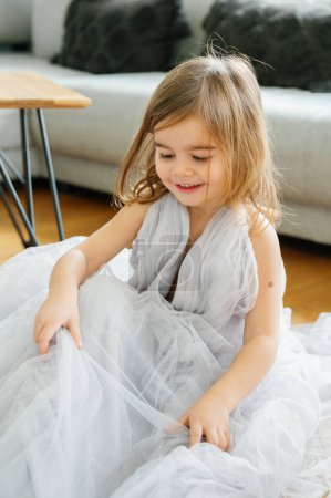 Téléchargez les photos : Une jeune fille gaie 3-4 ans portant une robe en tulle, aime jouer, enveloppé dans une robe en tulle doux à l'intérieur d'un salon confortable. - en image libre de droit