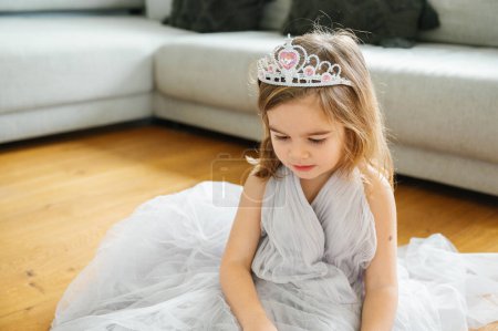 Téléchargez les photos : Une adorable fille sourit dans une robe en tulle, avec une tiare, assise sur le sol, portant une longue robe en tulle gris joie et des vibrations de princesse à la maison - en image libre de droit