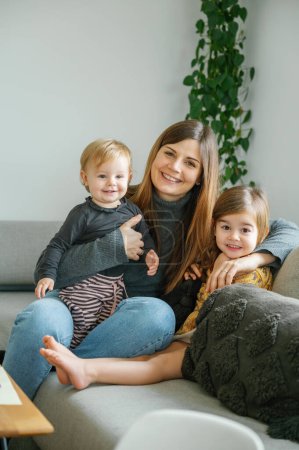 Foto de Una joven madre comparte un momento tierno con sus dos hijos, abrazados en un sofá en la sala de estar. Sus brazos amorosamente envuelven a un niño pequeño y a una hermana mayor. día relajado y alegre pasado en casa - Imagen libre de derechos