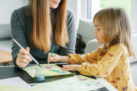 Photo for Crop of a kindergarten girl sitting at table, painting with watercolor and brush, a joyful art session unfolds as a mother helps her daughter with watercolor painting, fostering a love for art - Royalty Free Image