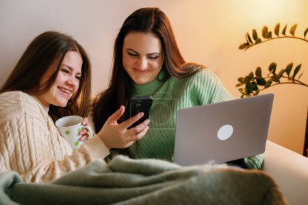 Two happy young women wearing jeans and sweat shirt, sitting on comfortable sofa with pillows, sharing fun moment while shopping online using smartphone and laptop on a cloudy day, business concept