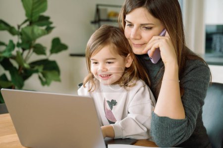 Téléchargez les photos : Une jeune fille souriante regarde un écran d'ordinateur pendant que sa mère parle au téléphone, gérant le travail à distance avec la vie de famille. - en image libre de droit