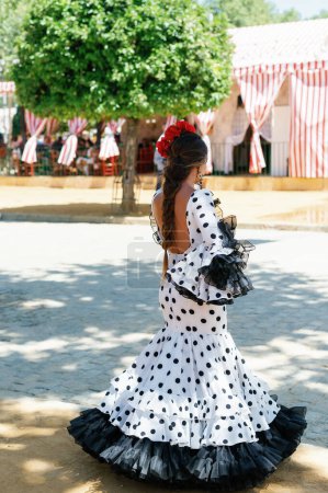 Photo for Back view of Beautiful woman wearing white flamenco dress with black dots. Detail of Traditional Spanish Hairstyle with red carnations. April Fair Feria de Abril, Seville Fair Feria de Sevilla - Royalty Free Image