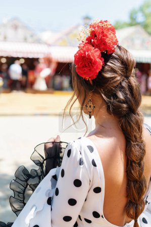 Photo for Back view of Beautiful woman wearing white flamenco dress with black dots. Detail of Traditional Spanish Hairstyle with red carnations. April Fair Feria de Abril, Seville Fair Feria de Sevilla - Royalty Free Image