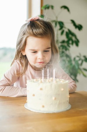 Foto de Una joven con la parte superior de color rosa pastel sentada a la mesa con una mirada enfocada apaga velas en un pastel de cumpleaños helado, un momento de delicia infantil y tradición de cumpleaños capturado en el interior - Imagen libre de derechos