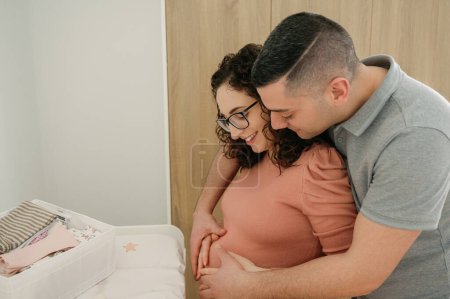 Foto de Joven feliz pareja embarazada, Hombre abrazando y tocando el vientre de su esposa embarazada en el futuro dormitorio del bebé, embarazo y concepto de personas - Imagen libre de derechos