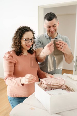 Téléchargez les photos : Photo verticale d'un jeune caucasien heureux couple enceinte pliant vêtements de bébé se préparer pour le jour de l'accouchement, la grossesse et le concept de famille - en image libre de droit