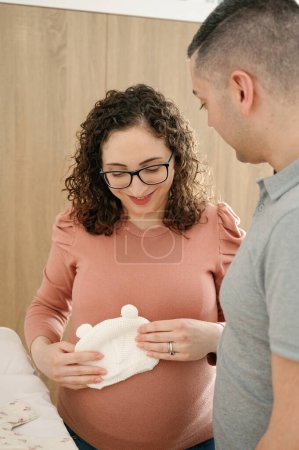 Foto vertical de una mujer embarazada feliz y su marido organizando ropa de bebé en la habitación, la familia y el concepto de embarazo