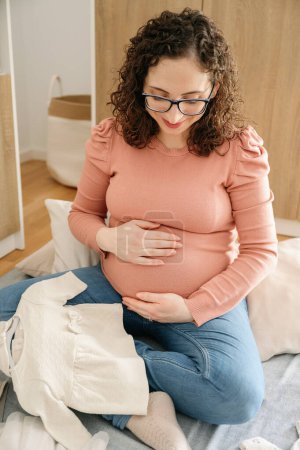 Téléchargez les photos : Photo verticale d'une femme enceinte occasionnelle de 30 à 35 ans, touchant son ventre assis sur la chambre à coucher pliant les vêtements de bébé. concept de famille - en image libre de droit