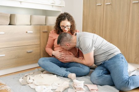 Téléchargez les photos : Homme touchant et embrassant le ventre de sa femme enceinte assise ensemble dans la chambre de bébé - en image libre de droit