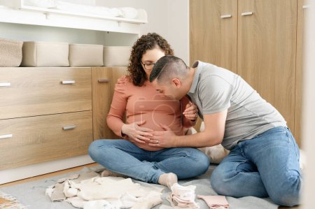 Photo for Man touching the belly of his pregnant wife, talking to baby girl in her belly, sitting on floor in the baby room. foreground baby clothes in background baby stuff, pregnancy and people concept - Royalty Free Image