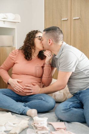 Téléchargez les photos : Photo verticale d'un couple enceinte tendre en vêtements décontractés s'embrassant sur la future chambre de bébé, homme touchant le ventre de sa femme enceinte, câlin. concept de famille - en image libre de droit