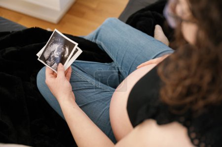 Photo for View from above the shoulder of a future mother looking at the baby ultrasound printed image wearing casual clothes, jeans and black top, sitting on the sofa. pregnancy concept - Royalty Free Image