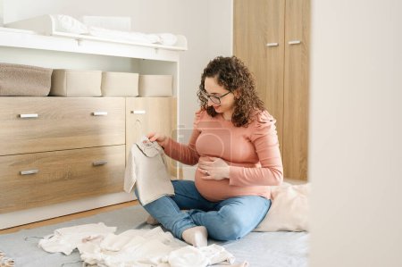 Téléchargez les photos : Heureux caucasien adulte enceinte femme préparant des vêtements d'accouchement assis sur le sol dans la chambre - en image libre de droit