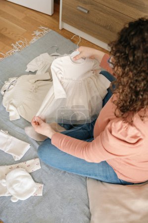 Téléchargez les photos : Vue de dessus verticale d'une femme enceinte adulte caucasienne méconnaissable se pliant et regardant les vêtements de bébé assis sur le sol dans la chambre - en image libre de droit