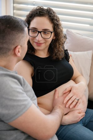Téléchargez les photos : Photo verticale d'un homme heureux touchant le ventre enceinte de sa femme assis sur le canapé près de la fenêtre à la maison, couple portant des vêtements décontractés - en image libre de droit