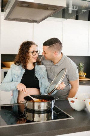 Téléchargez les photos : Photo verticale de deux heureux beaux amoureux enceintes homme et femme, se regardant tout en cuisinant à la maison, en se moquant. concept de famille - en image libre de droit