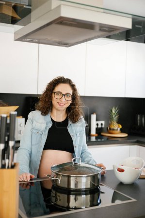 Photo for Vertical photo of a happy pregnant woman with brown curly hair, wearing eyeglasses standing in the kitchen smiling preparing food. food and family concept - Royalty Free Image