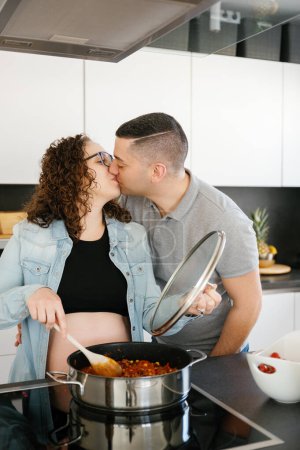 Téléchargez les photos : Photo verticale d'un couple enceinte embrassant pendant la cuisson dans la cuisine à la maison - en image libre de droit