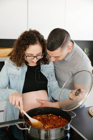 Téléchargez les photos : Photo verticale d'un homme touchant le ventre de sa femme enceinte pendant qu'elle cuisine à la maison, portant tous deux des vêtements décontractés, un haut, un jean, un t-shirt. concept de prengancy - en image libre de droit
