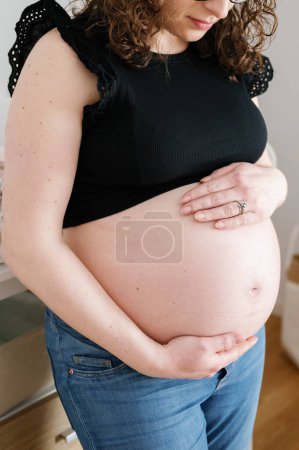Téléchargez les photos : Portrait vertical d'une femme enceinte debout les mains sur le ventre, portant un jean et un haut noir, regardant vers le ventre du bébé. maternité, personnes, concept d'attente - en image libre de droit