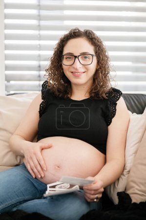 Téléchargez les photos : Portrait vertical d'une belle future mère heureuse avec un gros ventre sur le canapé, tenant une image échographique, regardant l'échographie - en image libre de droit