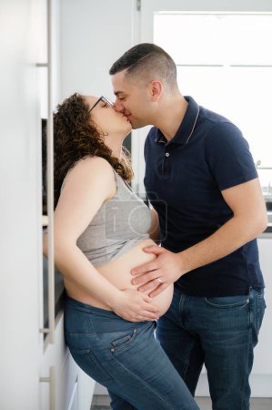 Téléchargez les photos : Vue latérale verticale portrait d'un couple enceinte embrassant joyeusement dans la cuisine - en image libre de droit