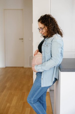 Téléchargez les photos : Vue latérale verticale portrait d'une femme enceinte mains sur le ventre debout dans le salon à la maison, grossesse, maternité, concept de personnes - en image libre de droit