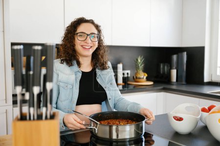 Téléchargez les photos : Portrait d'une femme enceinte heureuse souriante finissant de cuisiner, tenant un pot de nourriture faite maison dans les mains à la maison - en image libre de droit