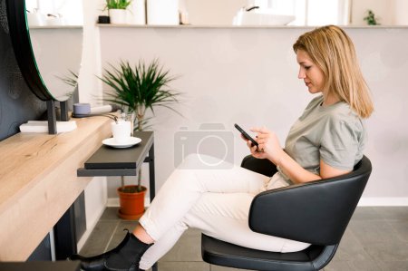 Téléchargez les photos : Vue latérale de la jeune belle femme assise dans une chaise dans un salon de beauté, en utilisant un téléphone mobile, se faire coiffer. concept beauté et soins capillaires - en image libre de droit
