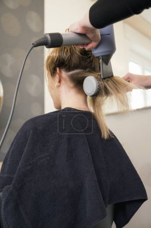 Photo for Back view of unrecognizable woman is getting her hair blow dried by a stylist. The stylist is using a round brush to style the womans hair - Royalty Free Image
