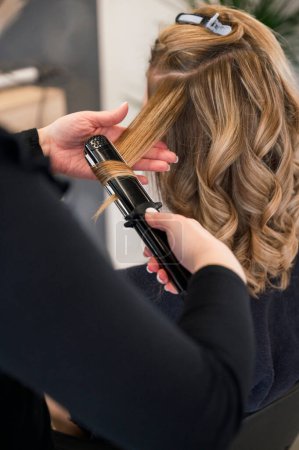 Téléchargez les photos : Vue arrière d'un styliste en utilisant un fer à friser pour coiffer les cheveux d'une femme. Le styliste porte une chemise noire et la femme une chemise noire. coiffeur et concept d'entreprise - en image libre de droit