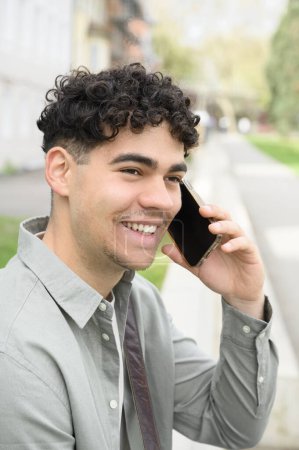 Téléchargez les photos : Jeune homme heureux en vêtements décontractés debout dans la rue et passer un appel téléphonique. concept de personnes - en image libre de droit